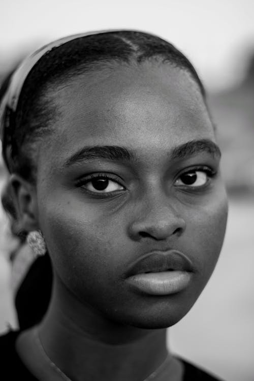 Black and white of feminine young African American lady with dark hair standing on street and looking at camera thoughtfully in daytime