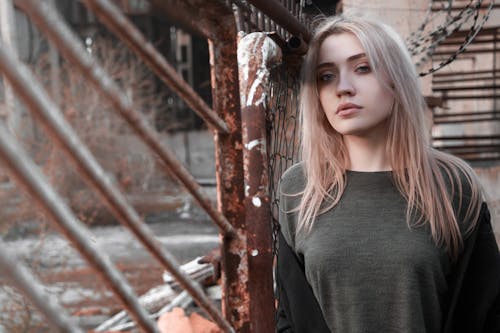 Young contemplative female in casual gray outfit leaning on grunge rusted metal fence in abandoned industrial building and looking at camera pensively