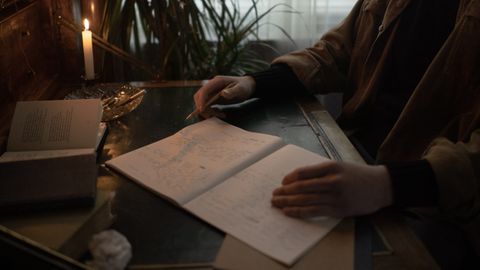 A person writes in a dimly lit room, surrounded by open books and candlelight, creating a moody study atmosphere.