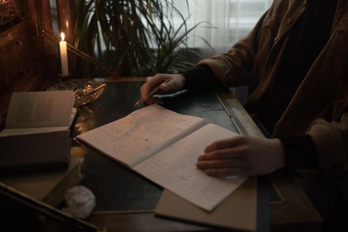 Free A person writes in a dimly lit room, surrounded by open books and candlelight, creating a moody study atmosphere. Stock Photo