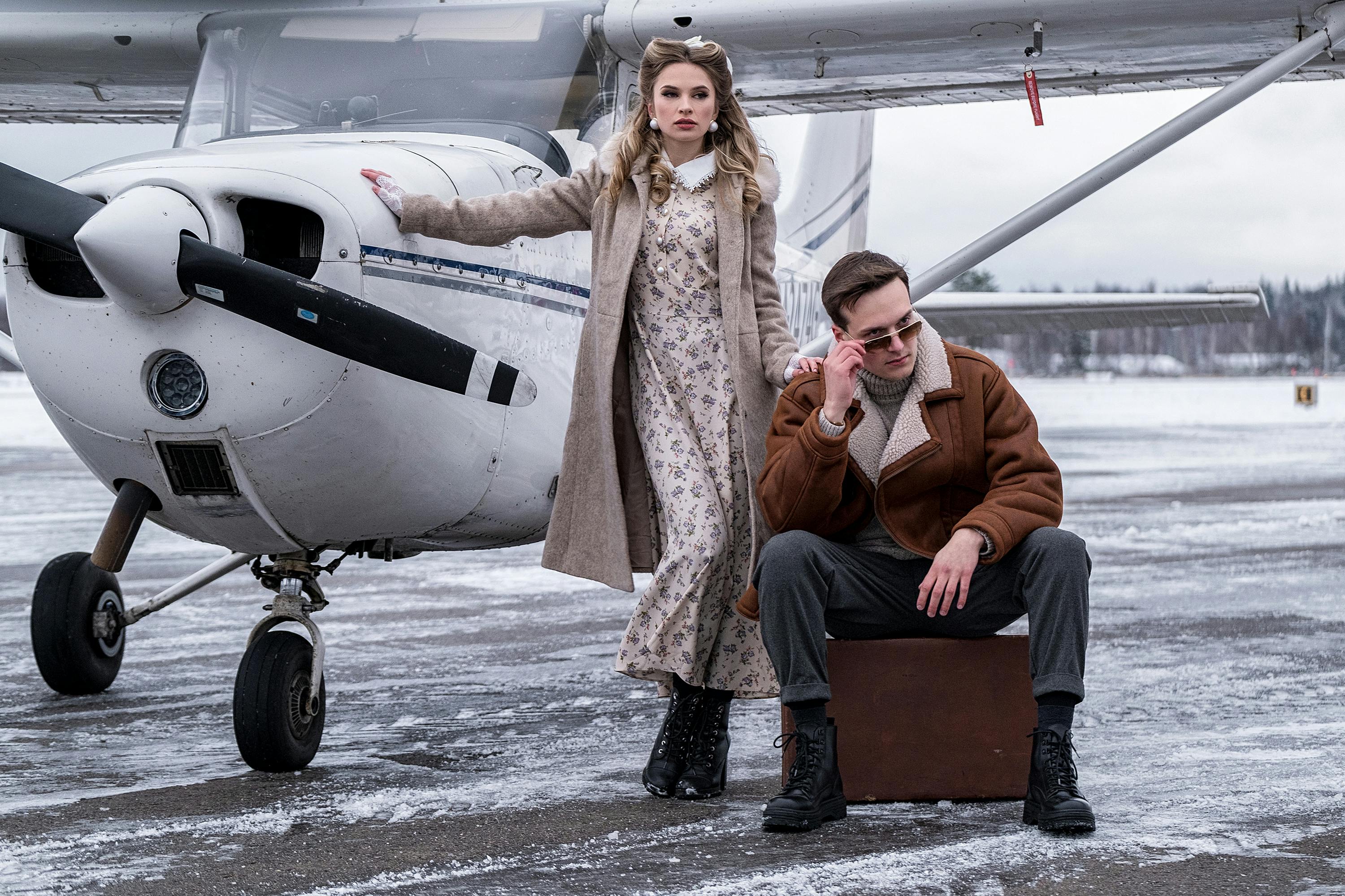 Full body trendy young couple in stylish warm wear standing near turboprop aircraft on snowy airfield