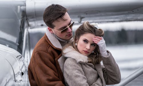 Side view young stylish couple in outerwear cuddling gently near modern propeller jet on snowy airfield