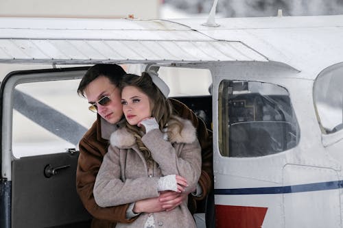 Stylish young couple in warm coats hugging gently near modern private jet and looking away on cold winter day