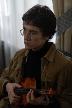 A young man in glasses and brown jacket playing the violin indoors, focused expression.