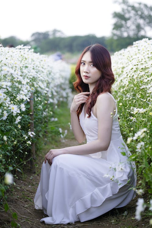 Charming Asian woman sitting near blooming flowers in countryside
