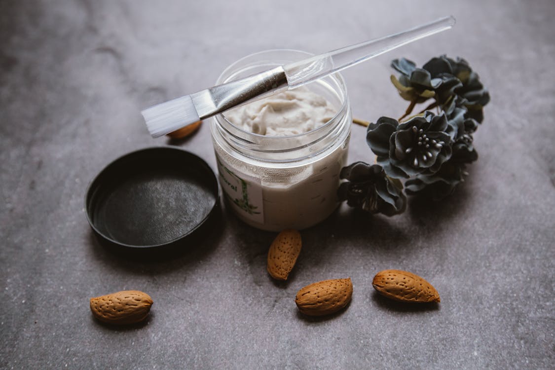 Body care mask with brush placed on table near scattered almonds and plant