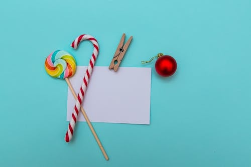Top view layout of striped candy cane and colorful lollipop composed with red Christmas bauble and white greeting card with paper clip on blue table