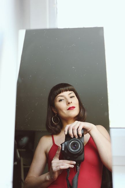 Woman in red top takes a selfie with a DSLR camera, reflecting in mirror.