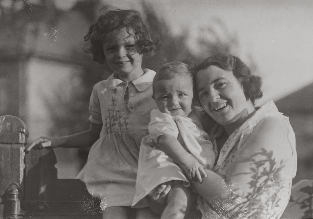 Una foto antigua en blanco y negro de una madre y sus dos hijos sonriendo al aire libre.