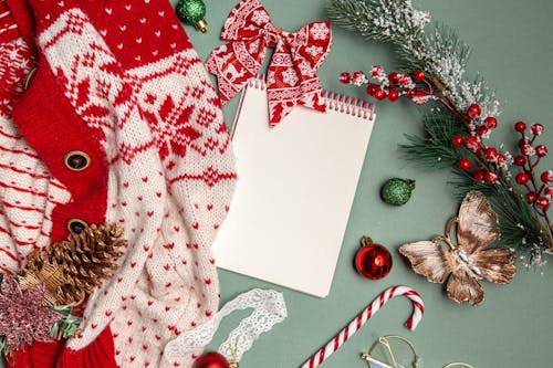 From above Christmas composition of stylish knitted cardigan and white notepad arranged with colorful baubles and fir cones and branch