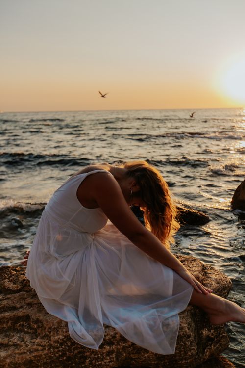 Free Side view of unrecognizable barefooted female traveler in white sundress embracing knees while relaxing on rocky seashore against cloudless sunset sky Stock Photo