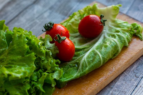 From above of assorted varieties of lettuce leaves placed on wet wooden cutting board with ripe appetizing tomatoes in kitchen in daylight