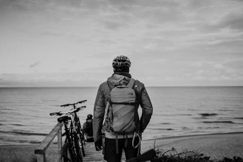 Black and white back view of anonymous male bicyclists with rucksacks and hand in pocket admiring ocean under cloudy sky
