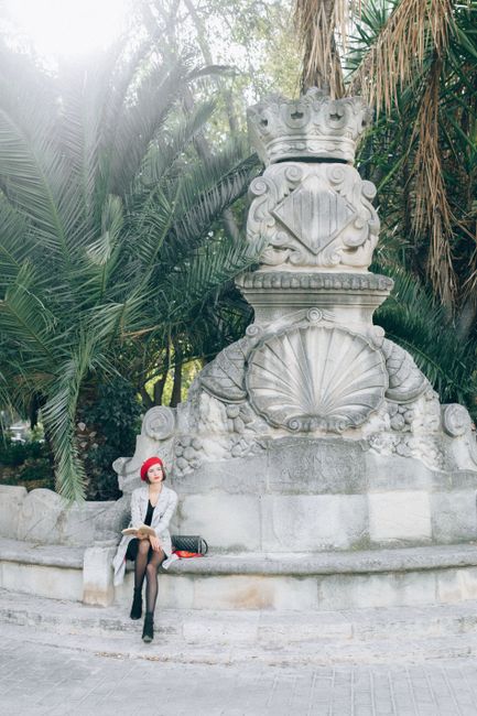 Stylish woman in a red beret sitting by a stone sculpture among palm trees.