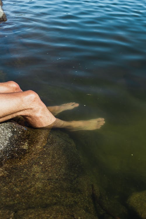 From above of crop faceless barefooted male traveler relaxing on rocky boulder with legs in lake water on sunny day