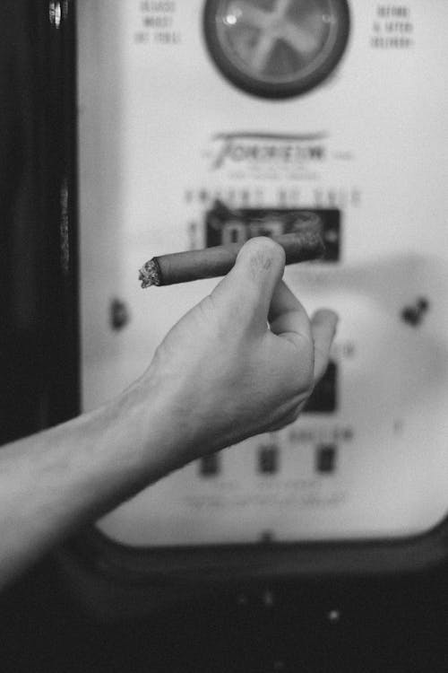 Black and white of crop unrecognizable male smoking cigar while standing on street near petrol machine