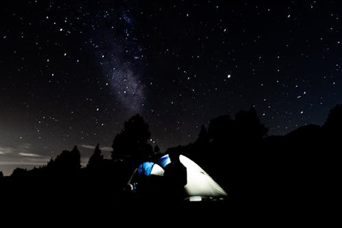 Silhouette of unrecognizable traveler standing near camp tent and observing majestic starry sky at dark night