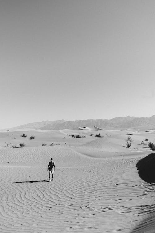 Black and white full body of anonymous male traveler resting on sandy dunes in desert valley on sunny day