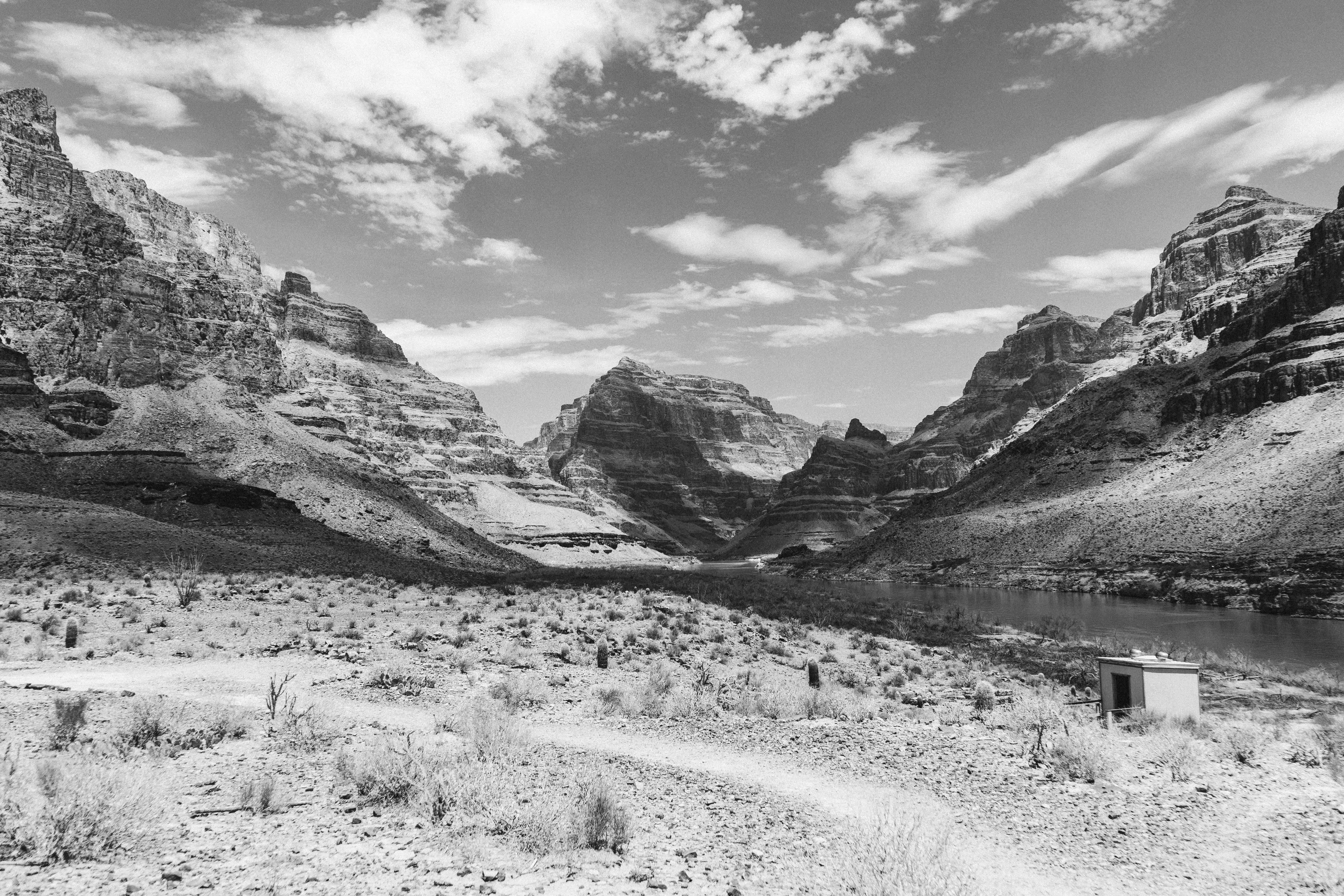 Picturesque black and white landscape of small cabin located near river flowing among rocky mountains in wild valley against cloudy sky