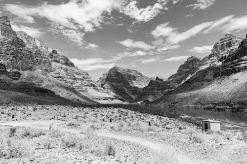 Picturesque black and white landscape of small cabin located near river flowing among rocky mountains in wild valley against cloudy sky