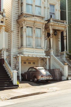 Retro car parked near aged white wooden house with staircases and ornamental details on windows located on city street on sunny day