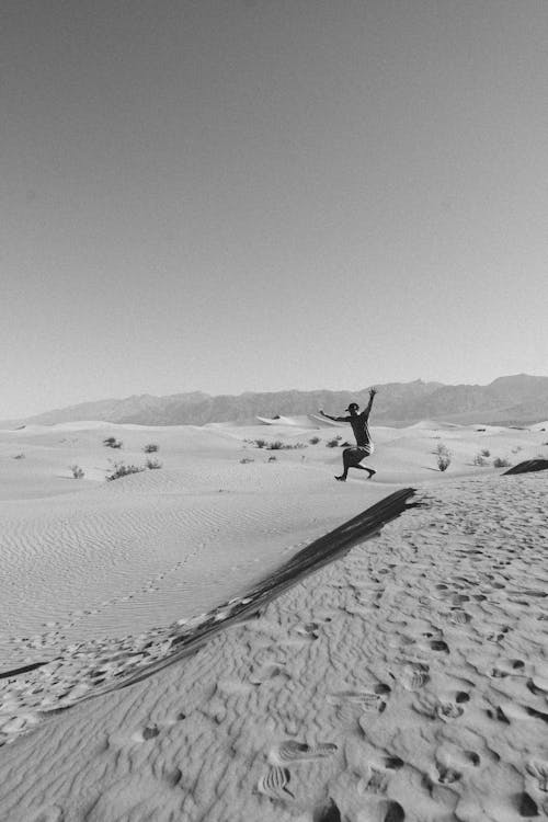 Black and white of unrecognizable male traveler jumping from sandy dune while having fun in desert on sunny day