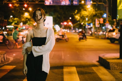 Young slender ethnic female looking at camera on blurred background of road with pedestrian crossing and lights in late evening