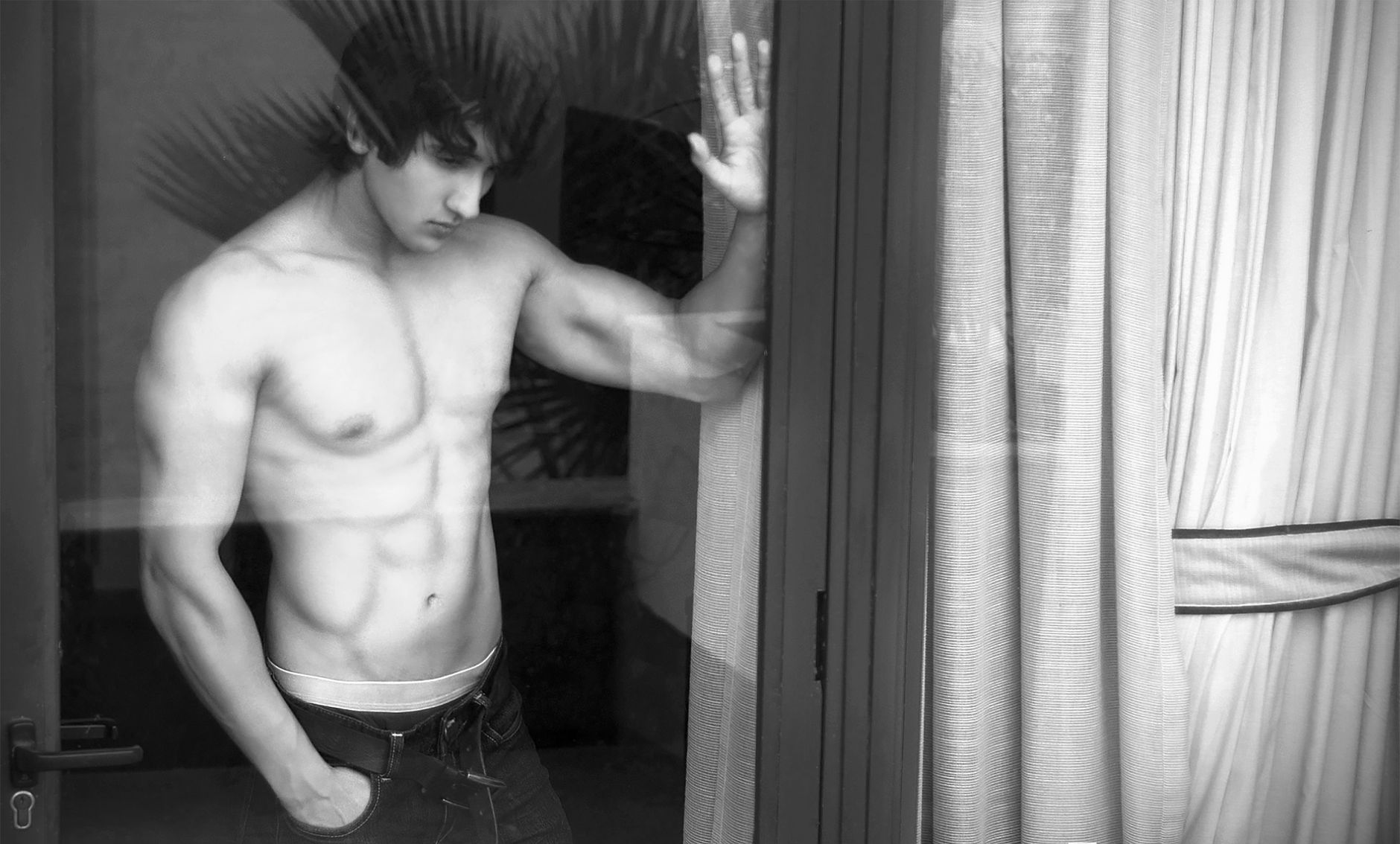 Black and white image featuring a shirtless male model with a muscular physique indoors.