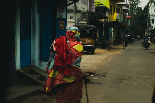Back view of unrecognizable ethnic indigenous person in rustic colorful clothes with hand stick walking on old shabby street in residential district