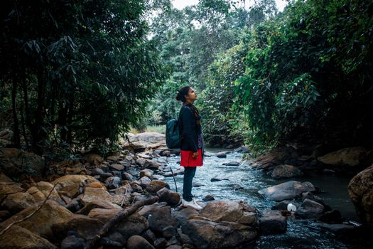 Side view of dreamy young ethnic female traveler in casual clothes and backpack standing on rocky coast of wild river flowing among green forest and enjoying nature