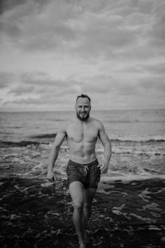 Black and white of young content shirtless fit traveler strolling on ocean coast and looking at camera