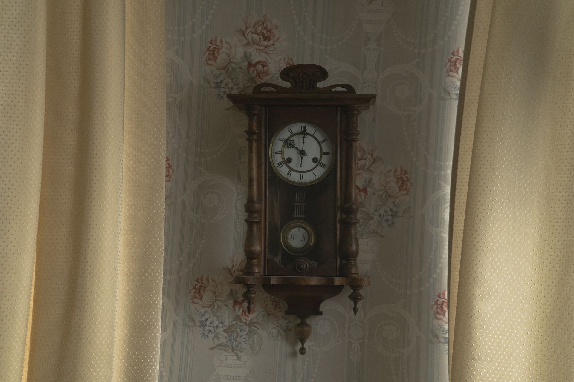 A vintage pendulum wall clock surrounded by floral wallpaper with curtains.