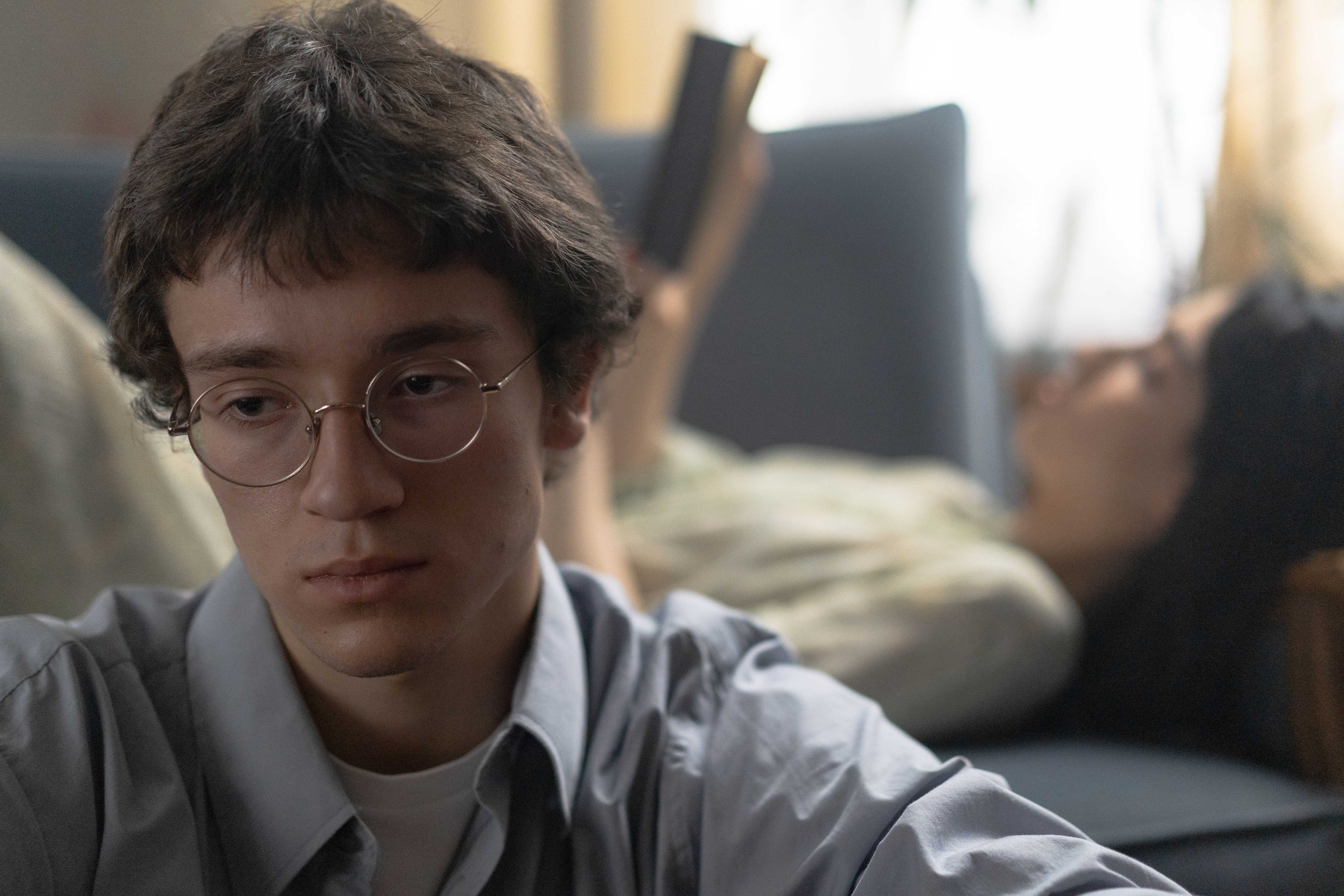 Young man in glasses looks thoughtful while a woman reads in the background, indoors.
