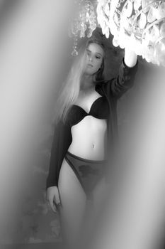 Black and white seductive young female with fit body wearing black underwear standing in light room and touching chandelier while looking at camera