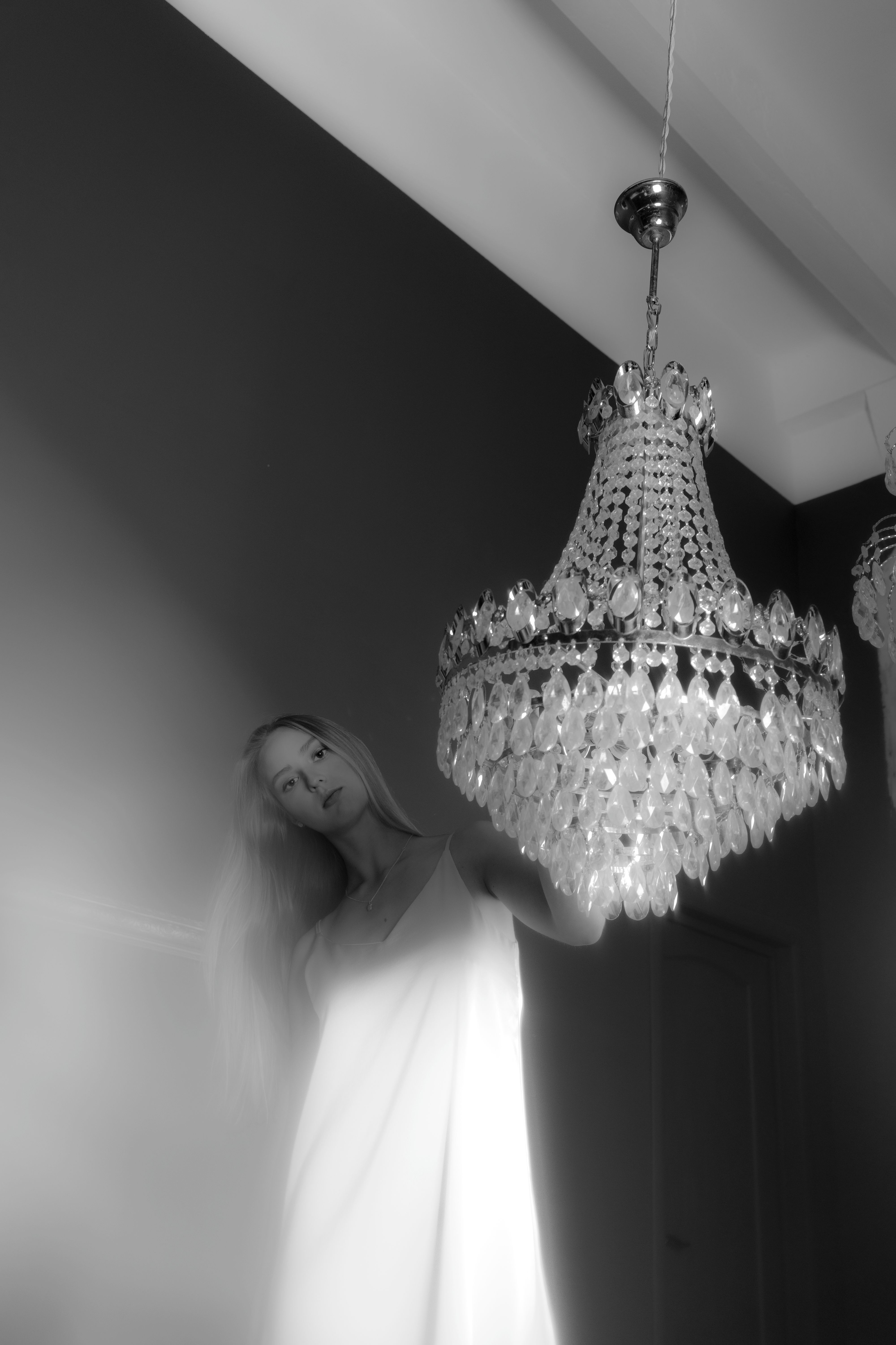 Black and white young female in white silk dress standing near hanging cut glass chandelier in light room