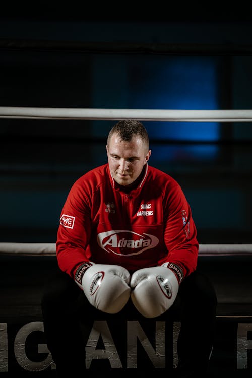 Powerful sportsman in sportswear and boxing gloves sitting in ring and looking down after practicing sport in gym