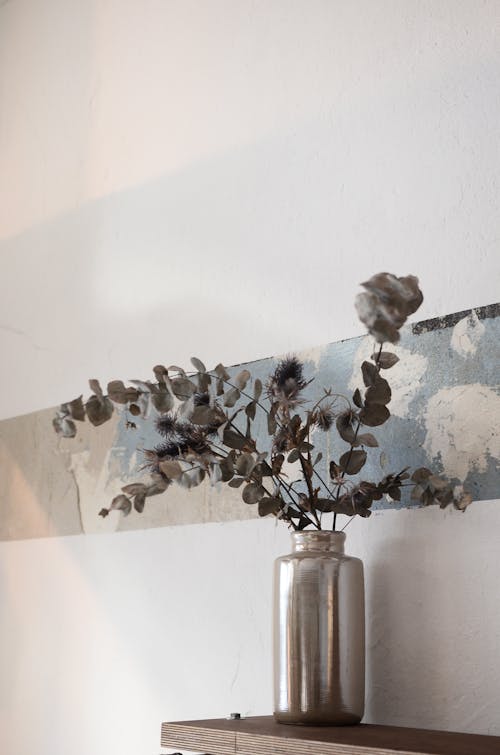 Shiny vase with dried plant sprigs with small leaves on wooden shelf near wall at home