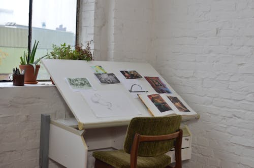Interior of art studio with different drawings and sketch on table near chair with potted plant and stationery on window