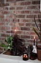 Green plants in pots near burning wax candles and carafe on cabinet against rough wall in house room