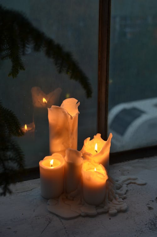 Burning candles with melted wax near plant leaf silhouette on windowsill at home in darkness