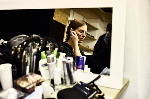 Reflection of female visagiste with brush applying makeup on face of model in dressing room