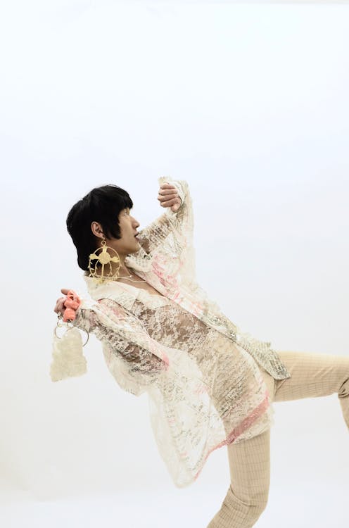 Side view of unusual Asian male in extravagant outfit with golden earrings dancing against white background