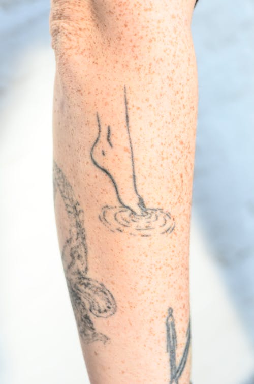 Free Crop unrecognizable person showing tattoos representing barefoot leg on tiptoe touching water on freckled skin Stock Photo