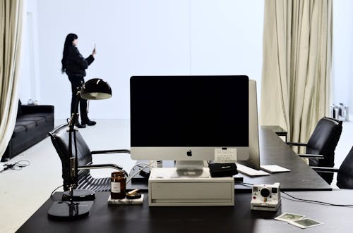 Monitor of modern computer and instant photo camera placed on black table in modern workspace against female standing with smartphone near window with light curtains