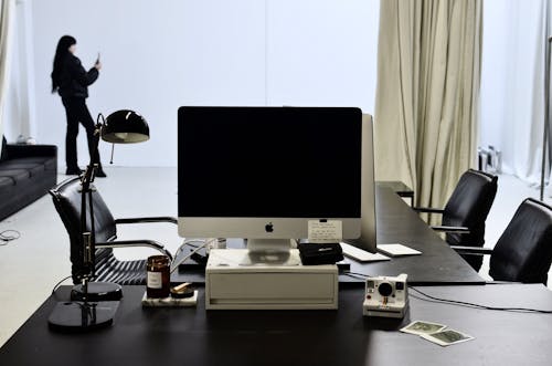 Contemporary computer placed on black table with instant photo camera and lamp against anonymous woman with long black hair in stylish clothes taking pictures on smartphone