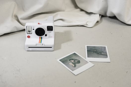 A white instant camera with rainbow stripe and two polaroid photos on a white floor.