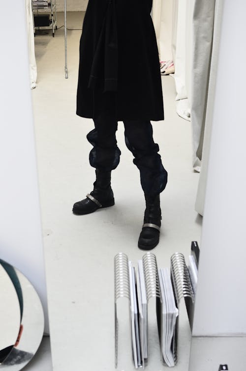 Crop anonymous person in loose trousers and black skirt reflecting in narrow mirror in light studio