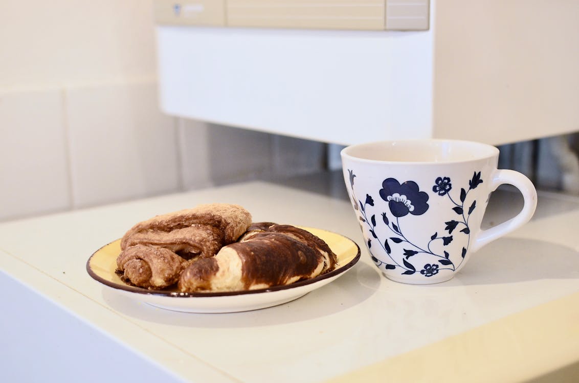 Mug Keramik Bunga Putih Dan Biru Disamping Roti Coklat Di Atas Piring Keramik Putih