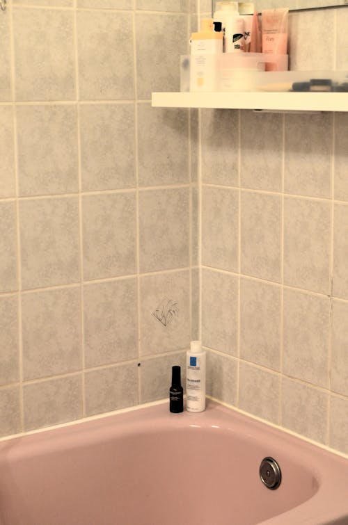 Plastic bottles with skincare products placed on edge of tub and shelf in bathroom with tiled wall during daily hygiene routine
