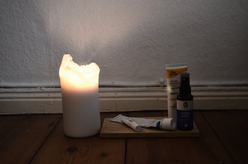White burning candle placed on wooden floor with vials and tubes for skincare daily routine against white shabby wall in room at low light